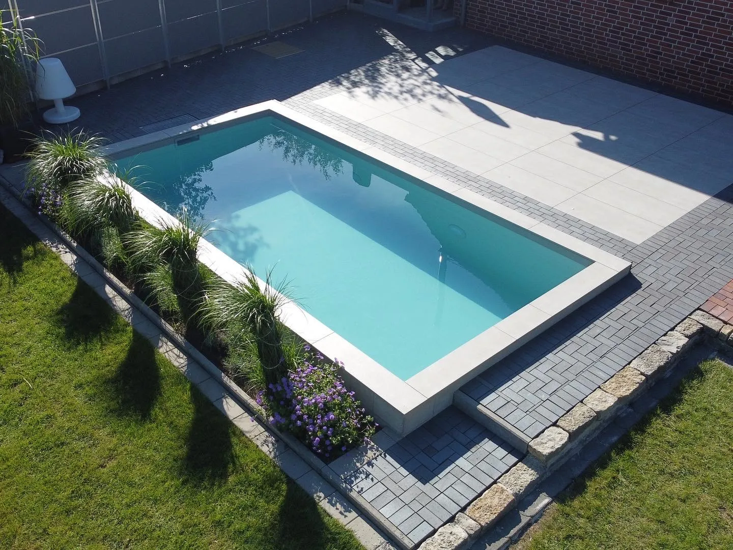 Poolgarten von oben: Klassisch eingefasst. Helle Umrandung, dunkler Belag, Rasen bis an die Kante.